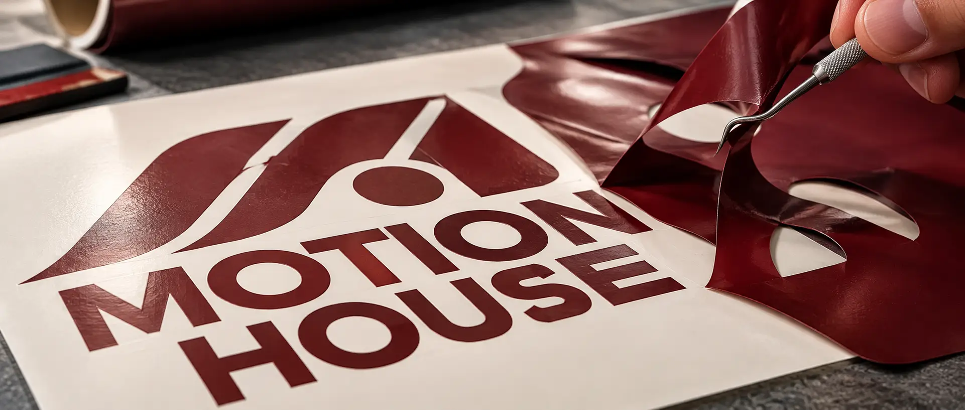 Uma ferramenta de descasque levanta o vinil vermelho-escuro excedente de um logotipo MOTION HOUSE cortado e revela os contornos de corte